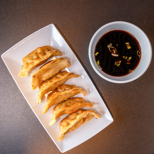 Seven golden-brown fried dumplings on a white rectangular plate with a side of soy sauce.